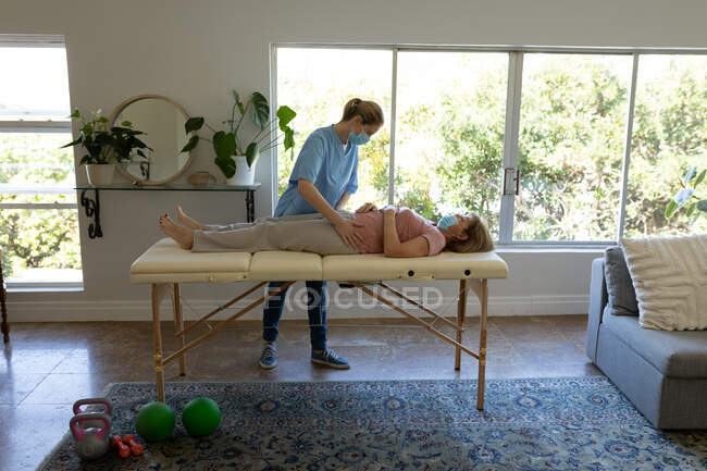 Senior Caucasian woman at home visited by Caucasian female nurse, stretching her hips, wearing face masks. Medical care at home during Covid 19 Coronavirus quarantine. — Stock Photo