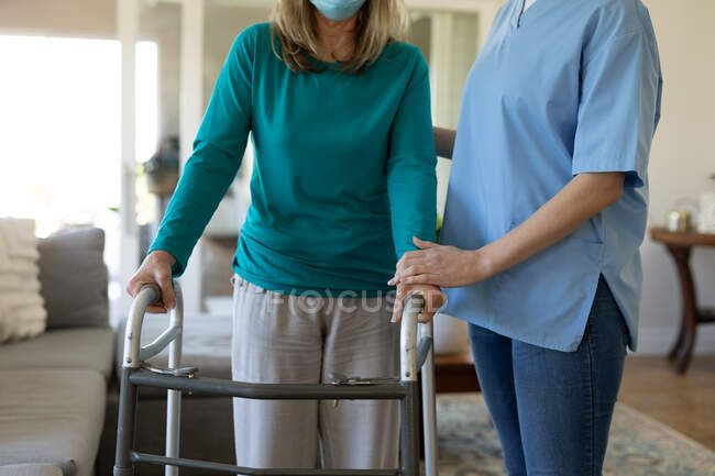 Eine ältere kaukasische Frau, die zu Hause von einer kaukasischen Krankenschwester besucht wird, geht mit einem Rollator und trägt eine Gesichtsmaske. Medizinische Versorgung zu Hause während der Quarantäne des Covid 19 Coronavirus. — Stockfoto