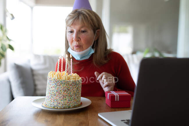 Старшая белая женщина проводит время дома, сидит в гостиной с праздничным тортом, в маске для лица и с ноутбуком. Социальное дистанцирование во время изоляции коронавируса Covid 19. — стоковое фото