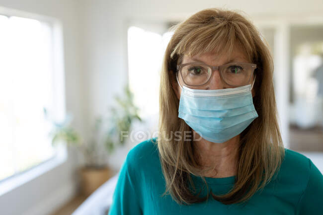 Portrait d'une femme caucasienne âgée passant du temps à la maison, debout dans son salon portant un masque facial. Distance sociale pendant le confinement en quarantaine du coronavirus Covid 19. — Photo de stock