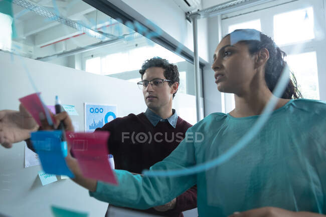 Mixed race woman and Caucasian man working in a casual office, writing on a glass board and talking. Creative business professionals working in a busy modern office. — Stock Photo