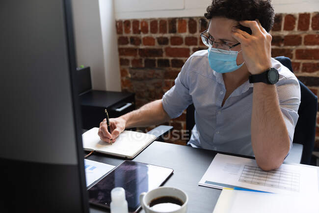 Uomo caucasico che lavora in un ufficio informale, prendendo appunti e indossando maschera facciale. Distanze sociali sul luogo di lavoro durante la pandemia di Coronavirus Covid 19. — Foto stock