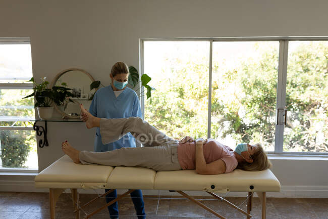 Mujer mayor caucásica en casa visitada por una enfermera caucásica, estirando la pierna, usando máscaras faciales. Coche médico - foto de stock
