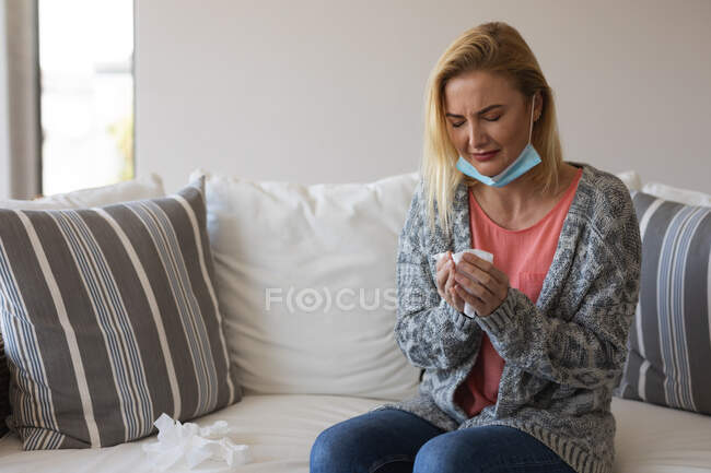 Белая женщина проводит время дома, в маске для лица, держит салфетку. Социальное дистанцирование во время изоляции коронавируса Covid 19. — стоковое фото