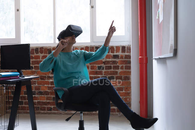Mixed race woman working in a casual office, wearing vr headset, touching virtual screen. Social distancing in the workplace during Coronavirus Covid 19 pandemic. — Stock Photo