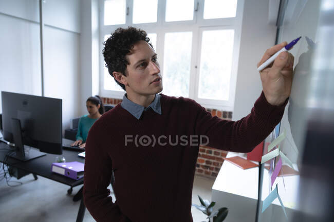 Caucasian man and mixed race woman working in a casual office, the man writing on a board, the woman using a computer. Social distancing in the workplace during Coronavirus Covid 19 pandemic. — Stock Photo