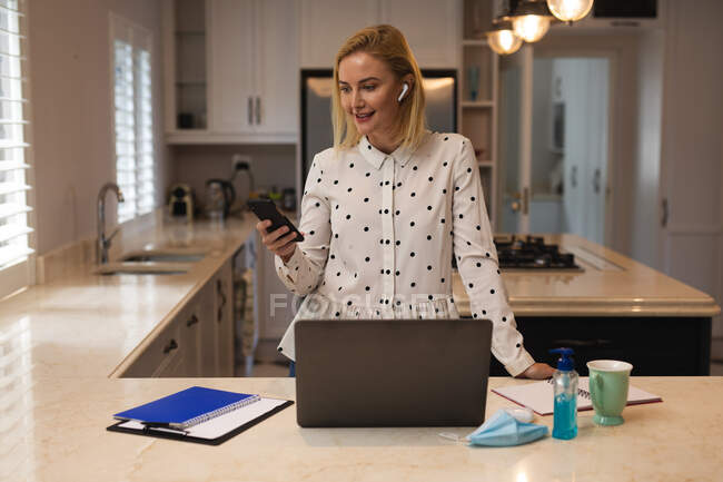Mulher branca trabalhando em casa, usando máscara facial, usando smartphone e laptop. Distanciamento social durante o bloqueio de quarentena do Covid 19 Coronavirus. — Fotografia de Stock