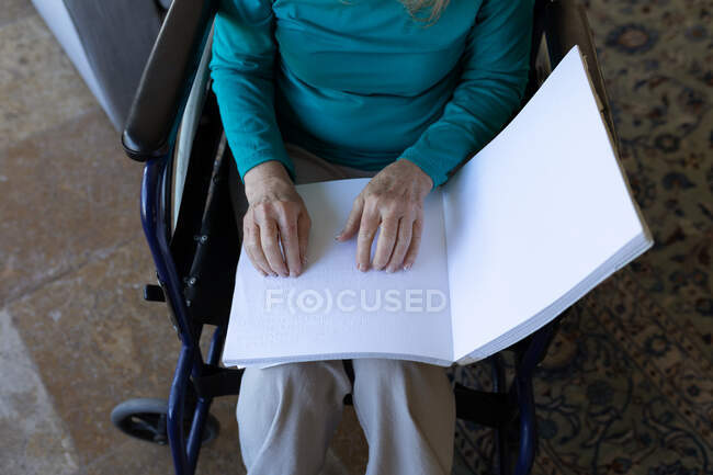 Женщина проводит время дома, сидя на инвалидной коляске и читая книгу руками. Социальное дистанцирование во время изоляции коронавируса Covid 19. — стоковое фото