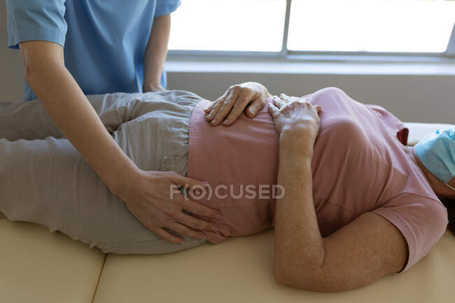 Mujer mayor caucásica en casa visitada por una enfermera caucásica, estirando sus caderas, usando mascarilla. Atención médica en el hogar durante la cuarentena del Coronavirus de Covid 19. - foto de stock