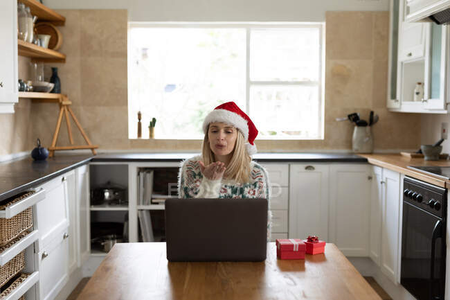 Kaukasische Frau verbringt Zeit zu Hause, sitzt zu Weihnachten mit Weihnachtsmütze in der Küche und benutzt Laptop mit Geschenken auf dem Tisch. Soziale Distanzierung während Covid 19 Coronavirus Quarantäne. — Stockfoto