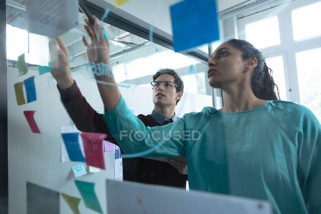 Femme métisse et homme caucasien travaillant dans un bureau décontracté, écrivant sur un panneau de verre et parlant. Professionnels créatifs d'affaires travaillant dans un bureau moderne occupé. — Photo de stock
