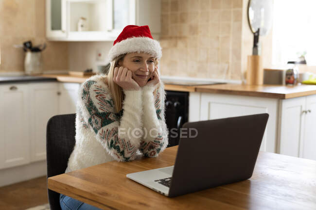 Caucasian woman spending time at home, sitting in kitchen at Christmas wearing Santa hat, using laptop. Social distancing during Covid 19 Coronavirus quarantine. — Stock Photo