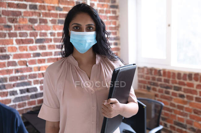 Портрет жінки змішаної раси, яка працює в повсякденному офісі, тримає ноутбук, носить маску обличчя і дивиться на камеру. Суспільна дистанція на робочому місці під час Коронавірусу Ковід 19 пандемії. — стокове фото