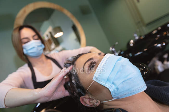 Белая женщина-парикмахер работает в парикмахерской в маске, моет волосы белой клиентки в маске. Здоровье и гиперактивность на рабочем месте во время коронавируса Ковид 19 пандемии. — стоковое фото