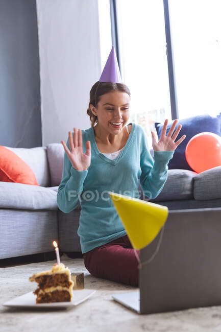 Happy Caucasian woman spending time at home, in party hat, sitting on floor using computer during video chat, birthday cake on floor. Social distancing during Covid 19 Coronavirus quarantine lockdown. — Stock Photo