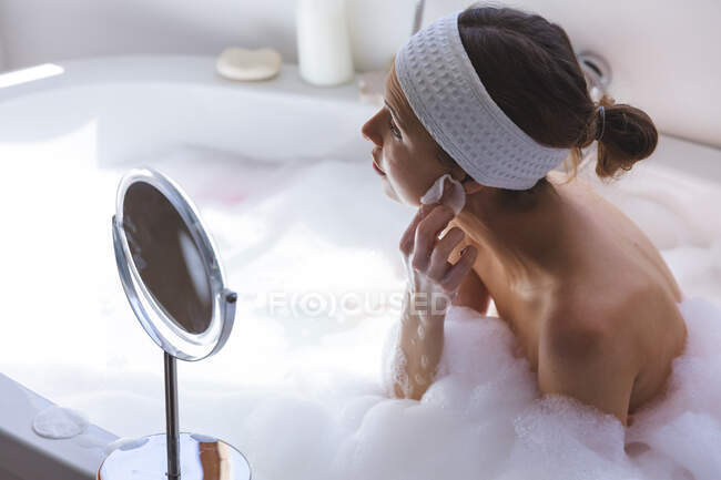 Mujer caucásica pasando tiempo en casa, en el baño, sentada en la bañera, mirando en el espejo limpiando su cara con almohadilla de algodón. Distanciamiento social durante el bloqueo de cuarentena del Coronavirus Covid 19. - foto de stock