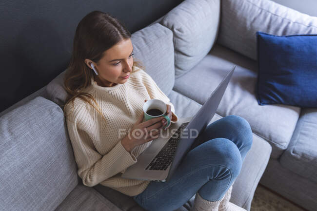 Donna caucasica trascorrere del tempo a casa, seduto sul divano in salotto utilizzando computer portatile con auricolari, tenendo tazza. Distanza sociale durante il blocco di quarantena Covid 19 Coronavirus. — Foto stock