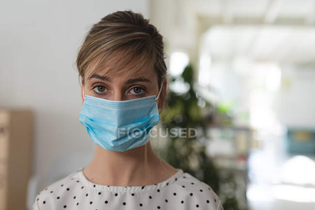 Retrato de negocio femenino caucásico creativo de pie en una oficina con máscara facial. Salud e higiene en el lugar de trabajo durante la pandemia de Coronavirus Covid 19. - foto de stock