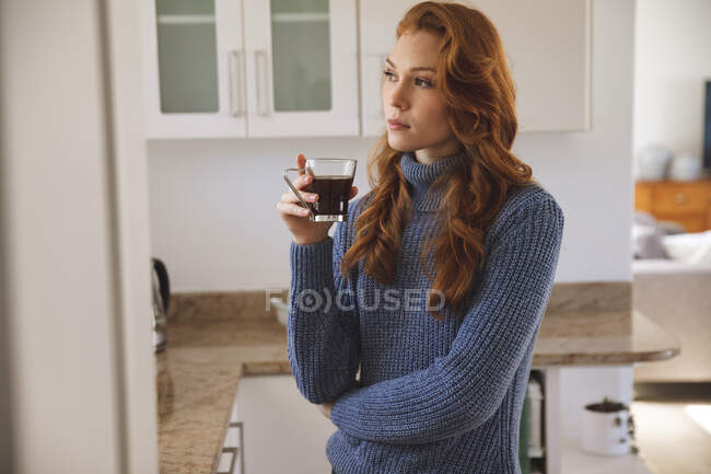 Mujer caucásica pasando tiempo en casa, en la cocina, mirando en serio, sosteniendo una taza, bebiendo café. Distanciamiento social durante el bloqueo de cuarentena del Coronavirus Covid 19. - foto de stock