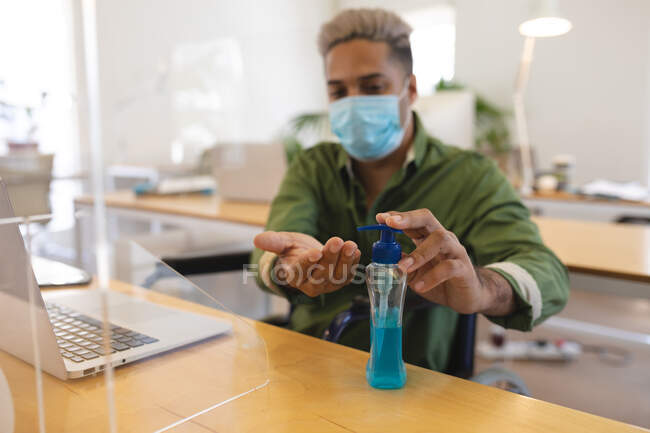 Mixed race male creative sitting at desk in an office wearing face mask disinfecting hands with hand sanitizer. Health and hygiene in workplace during Coronavirus Covid 19 pandemic. — Stock Photo