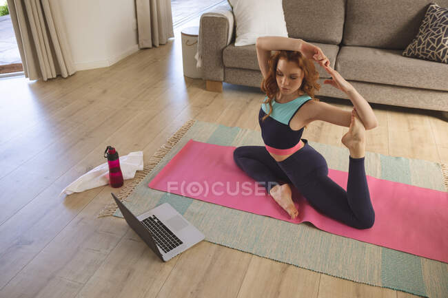 Кавказка проводит время дома, в гостиной, занимается спортом, практикует йогу, глядя на ноутбук. Социальное дистанцирование во время изоляции коронавируса Covid 19. — стоковое фото
