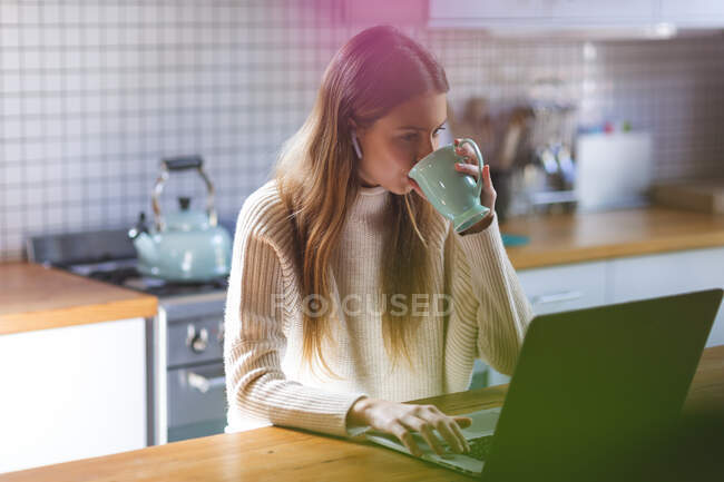 Donna caucasica trascorrere del tempo a casa, seduto in cucina utilizzando computer portatile con auricolari, bere da tazza verde. Distanza sociale durante il blocco di quarantena Covid 19 Coronavirus. — Foto stock