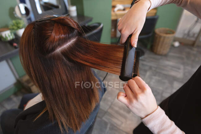 Hands of Caucasian female hairdresser working in hair salon wearing, straightening hair of female Caucasian customer. Health and hygiene in workplace during Coronavirus Covid 19 pandemic. — Stock Photo