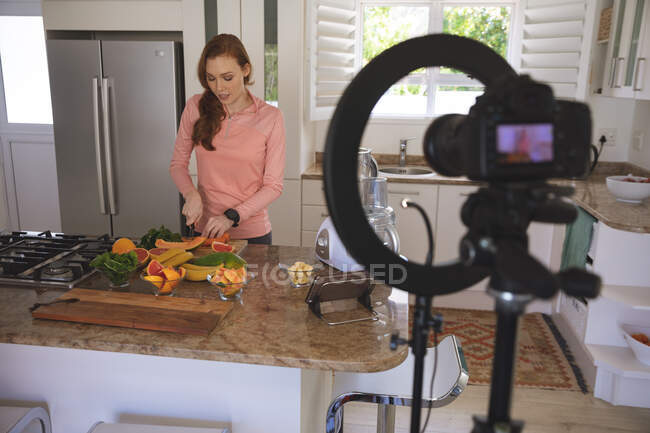 Белая женщина проводит время дома, режет фрукты на кухне, записывает их камерой. Социальное дистанцирование во время изоляции коронавируса Covid 19. — стоковое фото
