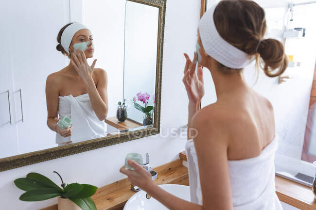 Caucasian woman spending time at home, standing in bathroom, looking in mirror applying face mask. Social distancing during Covid 19 Coronavirus quarantine lockdown. — Stock Photo