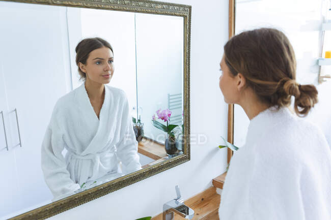Donna caucasica trascorrere del tempo a casa, in piedi in bagno, guardando nello specchio indossando accappatoio. Distanza sociale durante il blocco di quarantena Covid 19 Coronavirus. — Foto stock