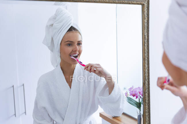 Femme caucasienne passant du temps à la maison, debout dans la salle de bain, regardant dans le miroir brossant les dents. Distance sociale pendant le confinement en quarantaine du coronavirus Covid 19. — Photo de stock
