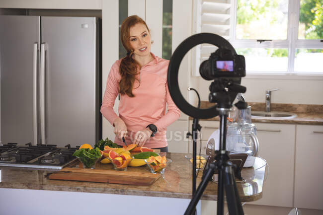 Белая женщина проводит время дома, режет фрукты на кухне, записывает их камерой. Социальное дистанцирование во время изоляции коронавируса Covid 19. — стоковое фото