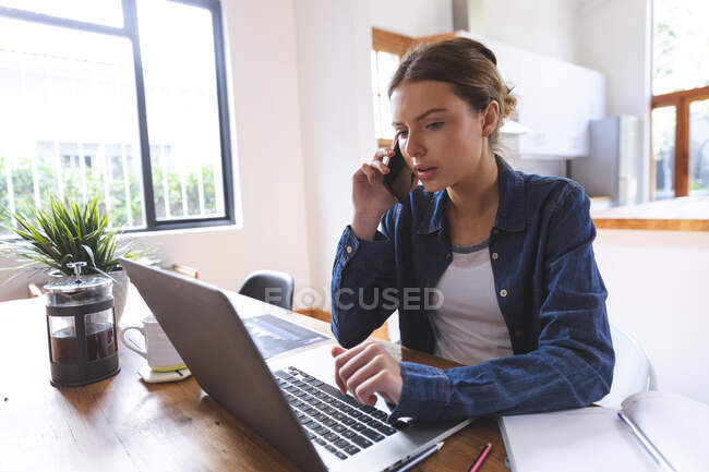 Mulher caucasiana passar o tempo em casa, sentado à mesa na cozinha usando computador portátil, trabalhando em casa, falando no smartphone. Distanciamento social durante o bloqueio de quarentena do Covid 19 Coronavirus. — Fotografia de Stock