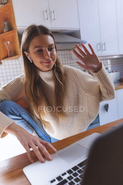 Caucasian woman spending time at home, sitting in kitchen using laptop computer with earphones, smiling and waving during video chat. Social distancing during Covid 19 Coronavirus quarantine lockdown. — Stock Photo