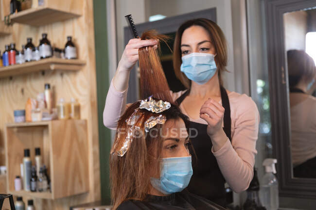 Caucasian female hairdresser working in hair salon wearing face mask, dying hair of female Caucasian customer in face mask. Health and hygiene in workplace during Coronavirus Covid 19 pandemic. — Stock Photo