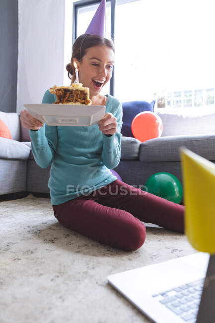 Happy Caucasian woman spending time at home, in party hat, sitting on floor using computer during video chat, holding birthday cake. Social distancing during Covid 19 Coronavirus quarantine lockdown. — Stock Photo