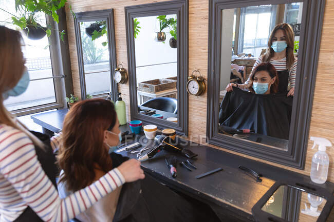 Белая женщина-парикмахер работает в парикмахерской в маске для лица, надевает накидку на белую клиентку в маске для лица. Здоровье и гиперактивность на рабочем месте во время коронавируса Ковид 19 пандемии. — стоковое фото
