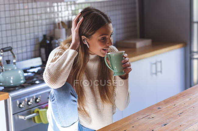 Mujer caucásica pasar tiempo en casa, sentado en la cocina con auriculares, sonriendo y sosteniendo taza verde. Distanciamiento social durante el bloqueo de cuarentena del Coronavirus Covid 19. - foto de stock