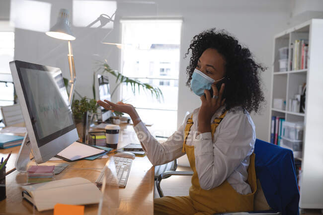 Смешанная расистка, работающая за столом в современном офисе в маске для лица и разговаривающая по смартфону. Здоровье и гипертония на рабочем месте во время коронавируса Ковид 19 пандемический. — стоковое фото