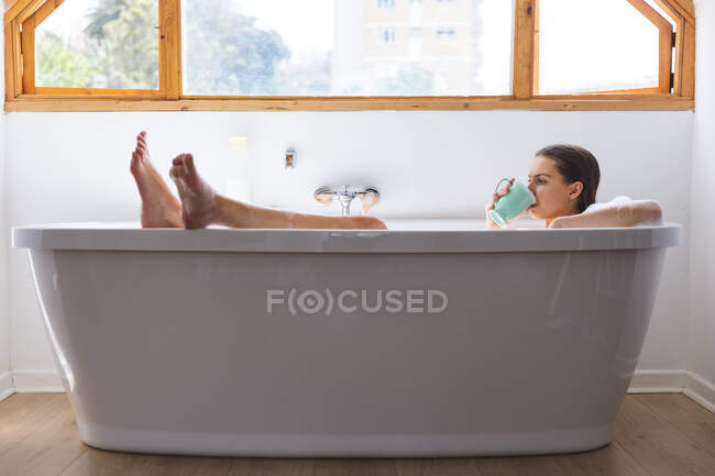 Кавказька жінка проводить час удома, у ванній кімнаті, лежачи в ванній, розслабившись від чашки. Соціальна дистанція в Ковиді 19 Коронавірус карантин. — стокове фото