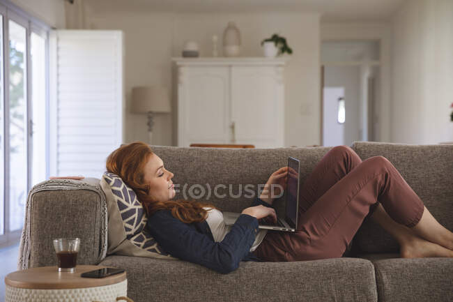 Mujer caucásica pasar tiempo en casa, en la sala de estar, utilizando el ordenador portátil, acostado en el sofá. Distanciamiento social durante el bloqueo de cuarentena del Coronavirus Covid 19. - foto de stock