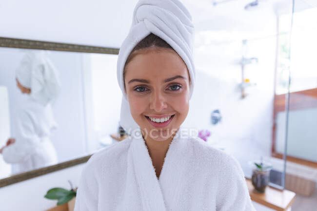 Retrato de una mujer caucásica feliz pasando tiempo en casa, de pie en el baño, sonriendo a la cámara con una toalla alrededor de su cabello. Distanciamiento social durante el bloqueo de cuarentena del Coronavirus Covid 19. - foto de stock