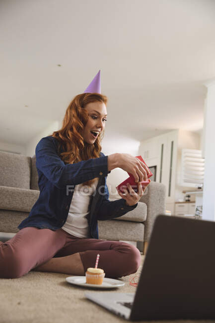 Mujer caucásica pasando tiempo en casa, en la sala de estar, sonriendo, celebrando, abriendo un regalo, magdalena a su lado. Distanciamiento social durante el bloqueo de cuarentena del Coronavirus Covid 19. - foto de stock