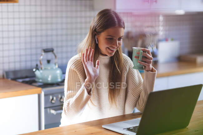 Mujer caucásica pasar tiempo en casa, sentado en la cocina con el ordenador portátil con auriculares, sonriendo y saludando durante el chat de vídeo. Distanciamiento social durante el bloqueo de cuarentena del Coronavirus Covid 19. - foto de stock