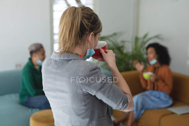 Multi ethnic group of male and female business creatives wearing face masks and distancing holding coffees and talking. Health and hygiene in workplace during Coronavirus Covid 19 pandemic. — Stock Photo