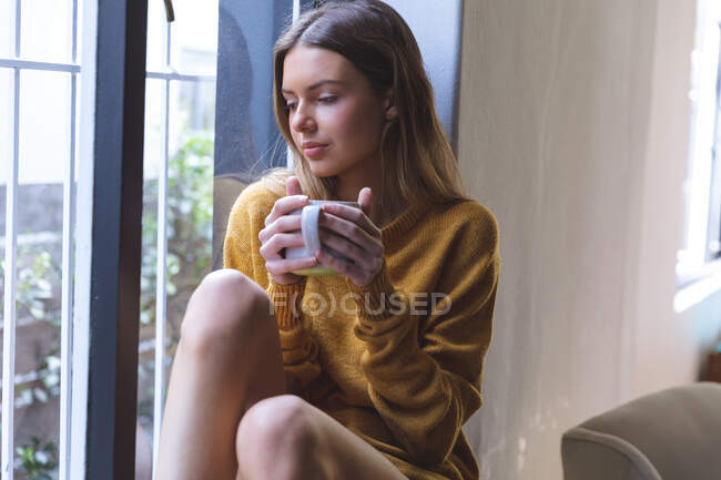 Femme caucasienne passant du temps à la maison, assise sur le rebord de la fenêtre dans le salon, tenant une tasse verte. Distance sociale pendant le confinement en quarantaine du coronavirus Covid 19. — Photo de stock