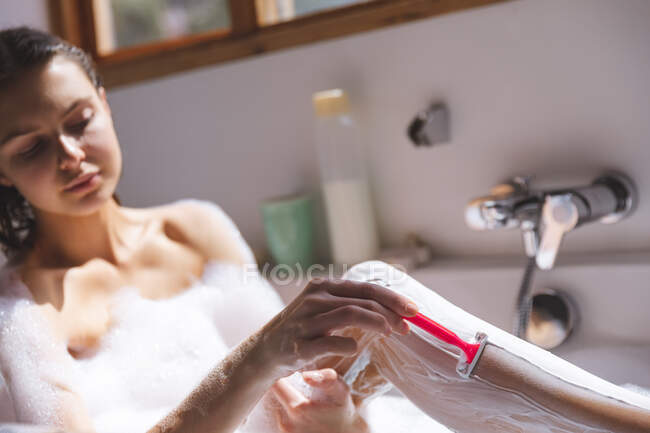 Mujer caucásica pasar tiempo en casa, en el baño, sentado en la bañera, piernas de afeitado. Distanciamiento social durante el bloqueo de cuarentena del Coronavirus Covid 19. - foto de stock