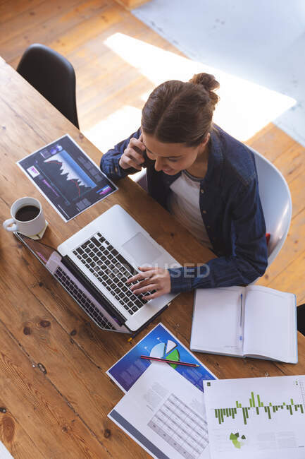 Caucasian woman spending time at home, sitting by table in kitchen using laptop computer, working from home, talking on smartphone. Social distancing during Covid 19 Coronavirus quarantine lockdown. — Stock Photo