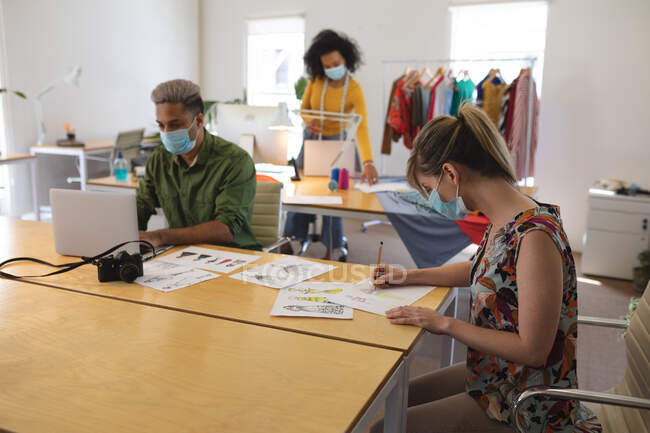 Multi ethnic group of male and female fashion designers working in studio wearing face masks and distancing. Health and hygiene in workplace during Coronavirus Covid 19 pandemic. — Stock Photo