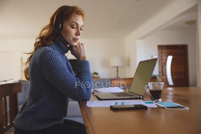 Mulher caucasiana passando tempo em casa, na cozinha, trabalhando em casa, usando seu laptop, usando um fone de ouvido. Distanciamento social durante o bloqueio de quarentena do Covid 19 Coronavirus. — Fotografia de Stock